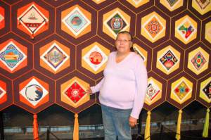 Marlene Shawanda stands next to her Ojibwe rose quilt block, representing the Ojibwe people of  Canada. The colours used in her depiction represent the Four Directions.