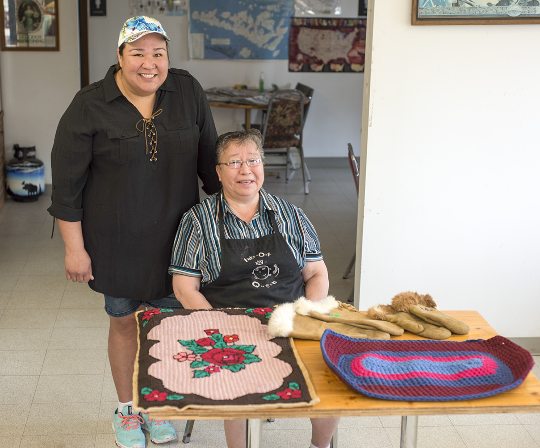 Betsy Debassige (nee Bebamash) and Holly display some of the handmade rugs and moose hide gloves that Maggie made. The rugs are made from old t-shirts and are and woven onto a potato sack backing.