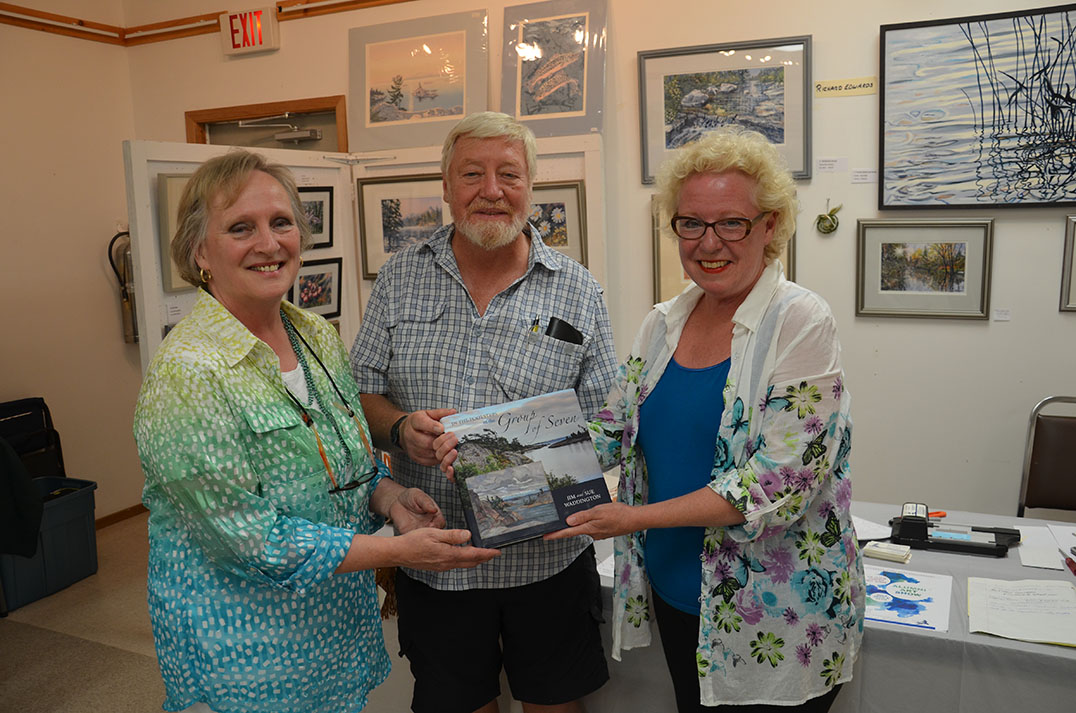 Instructor Richard Edwards and volunteer/artist Sylvia Simpson present longtime organizer Ellen Lewis with a copy of ‘In the Footsteps of the Group of Seven’ signed by the alumni of the LaCloche Art School. photos by Michael Erskine