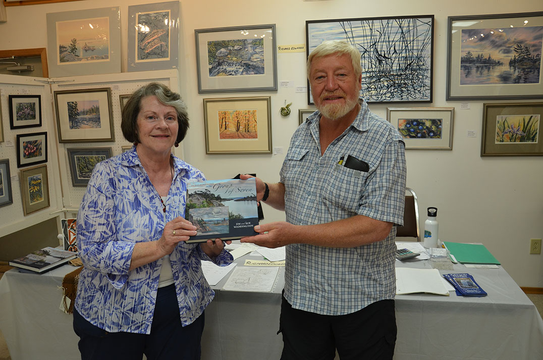 Volunteer/artist Pauline Toffelmyer of Sheguiandah presents instructor Richard Edwards of Kagawong with a copy of ‘In the Footsteps of the Group of Seven’ signed by all of the alumni. So secret were the presentation plans even Mr. Edwards inadvertently signed his copy.