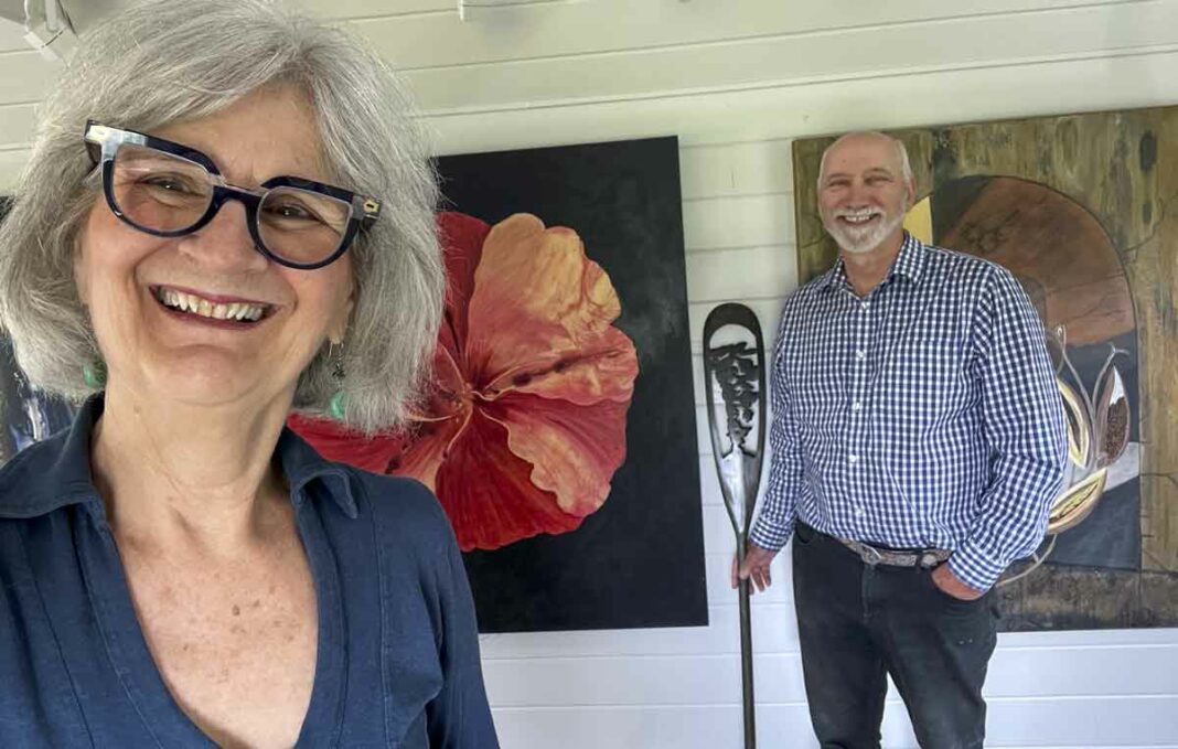 Audrey Jolly and Ed Horner are all smiles while posing for a photograph in their art studio. Ms. Jolly is a multi-discipline artist who works in a host of media and is an accomplished artists and author while her pparnter Mr. Horner is quite the carver. He is seen here holding one of his intricately carved paddles with Ms. Jolly’s artworks in the background.