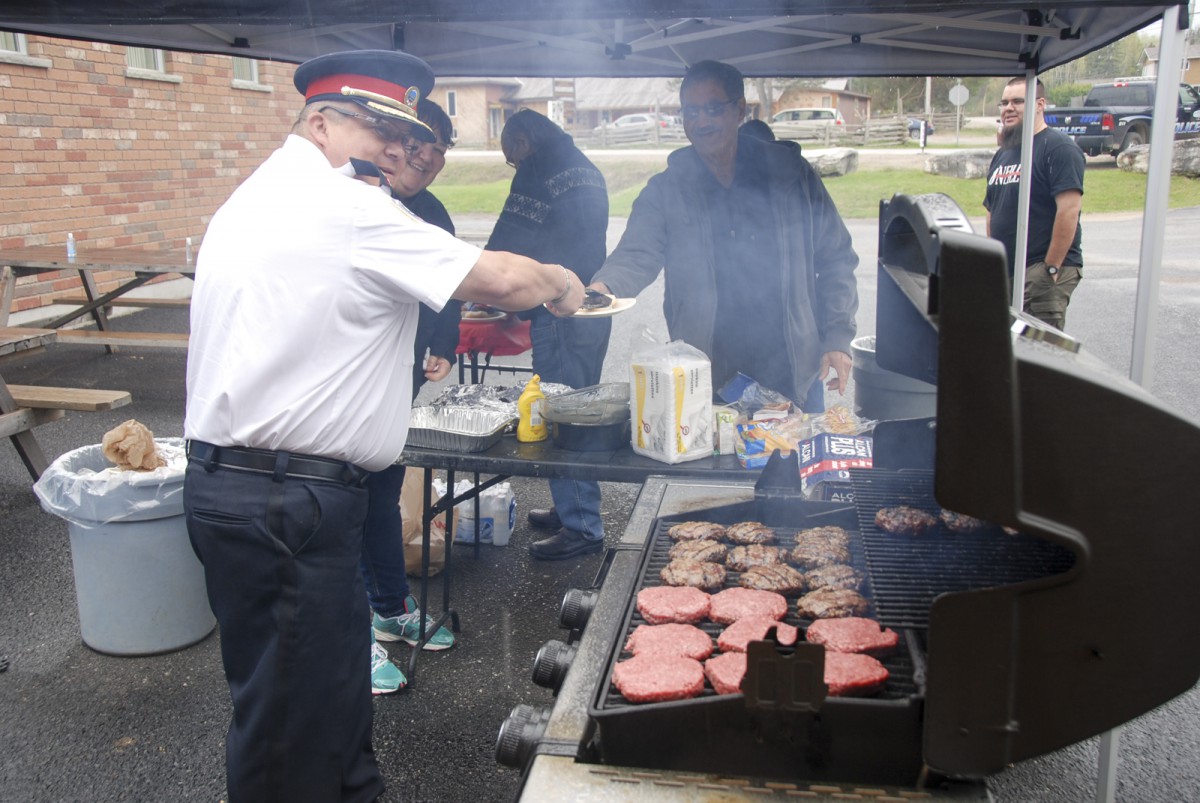 UCCM Police Chief Rodney Nahwegahbow serves up a burger  during the force’s 20th anniversary celebrations while front desk staffer Ann Ziegler keeps a close eye on his work.