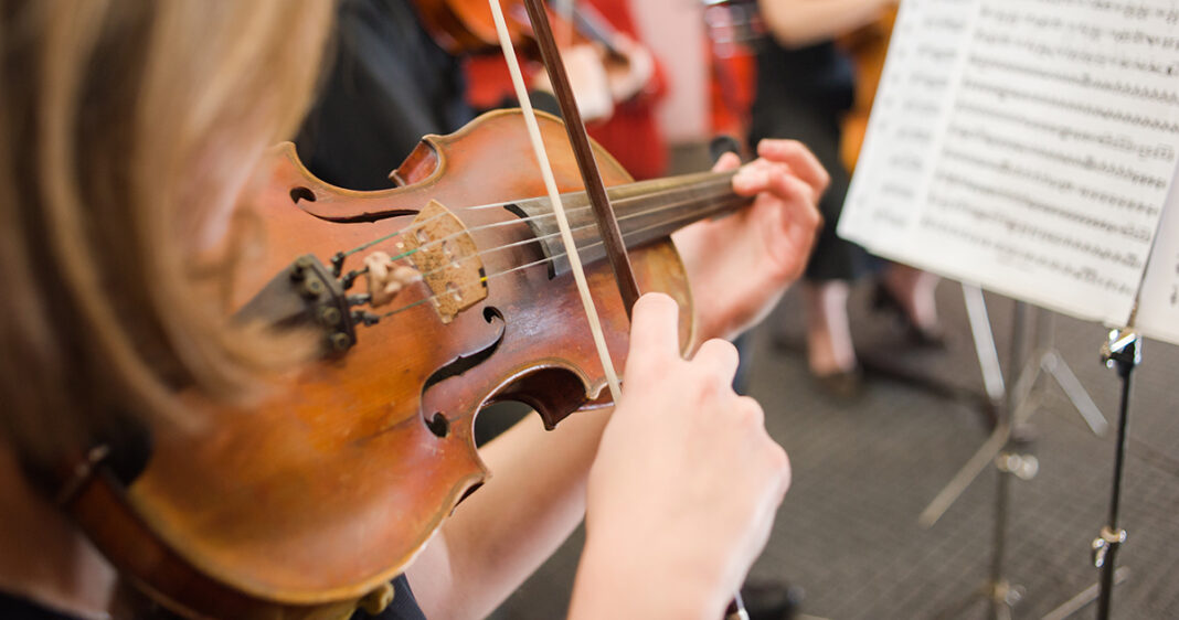 violin being played and sheet music in foreground with other musicians in background